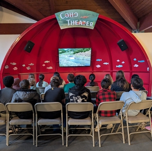 Elementary students sitting in chairs in front of the Coho Theater installation inside the fish hatchery, watching a video about the steelhead life cycle.
